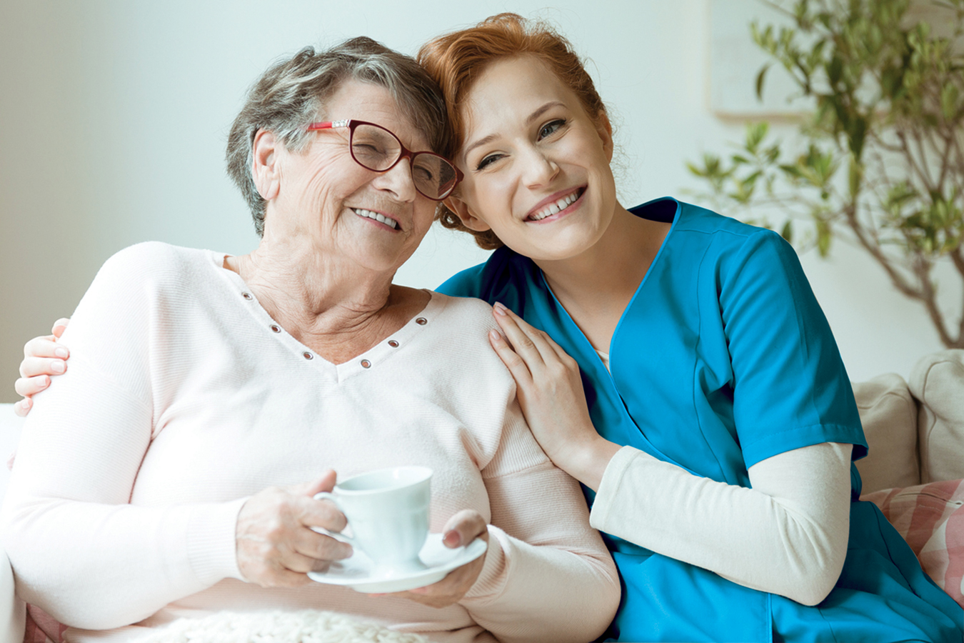 Smiling nurse in beige uniform hugs old lady drinking tea while sitting on white sofa in nursing home