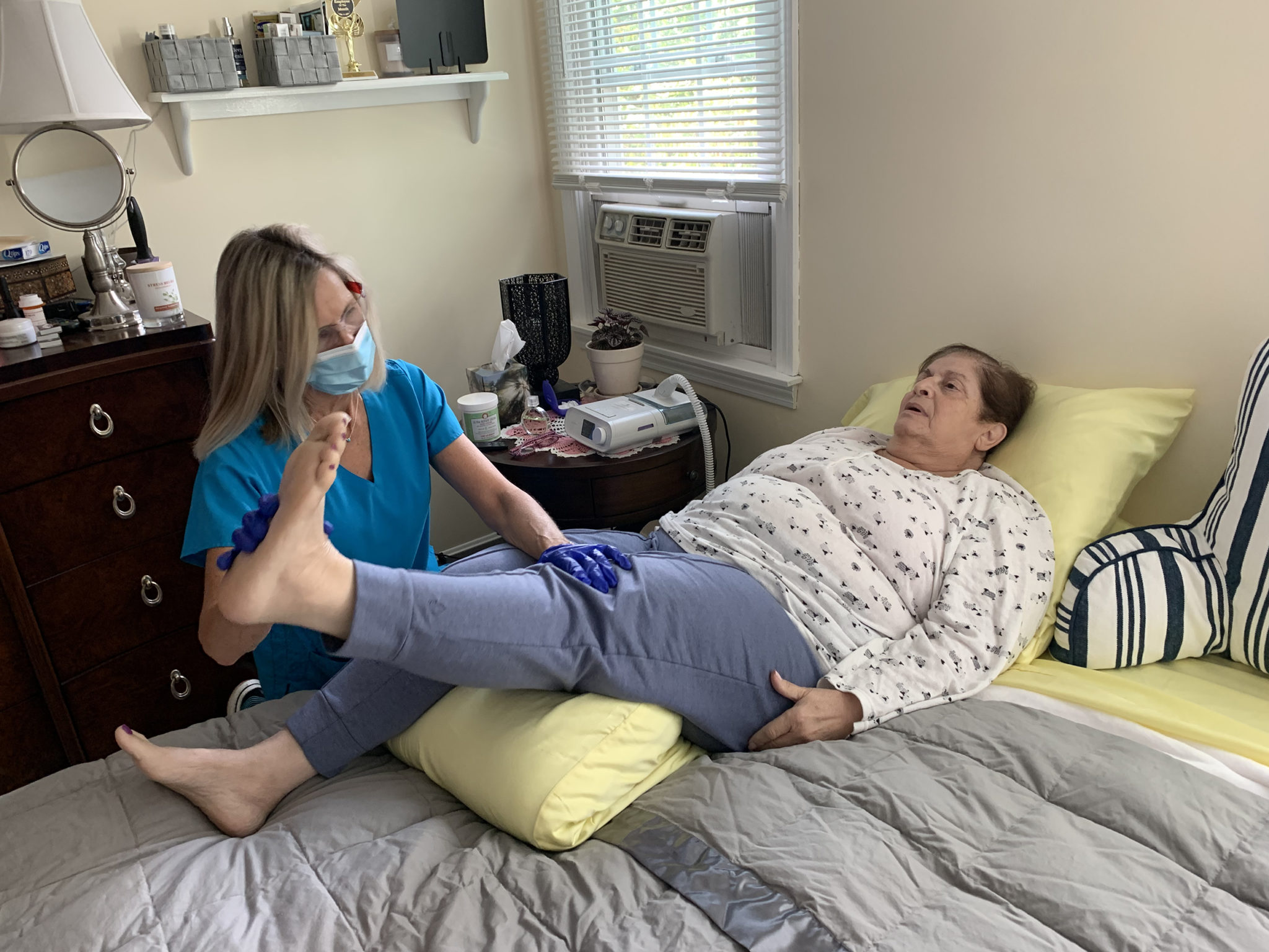 Woman in teal scrubs help older woman lying in bed do leg lifts