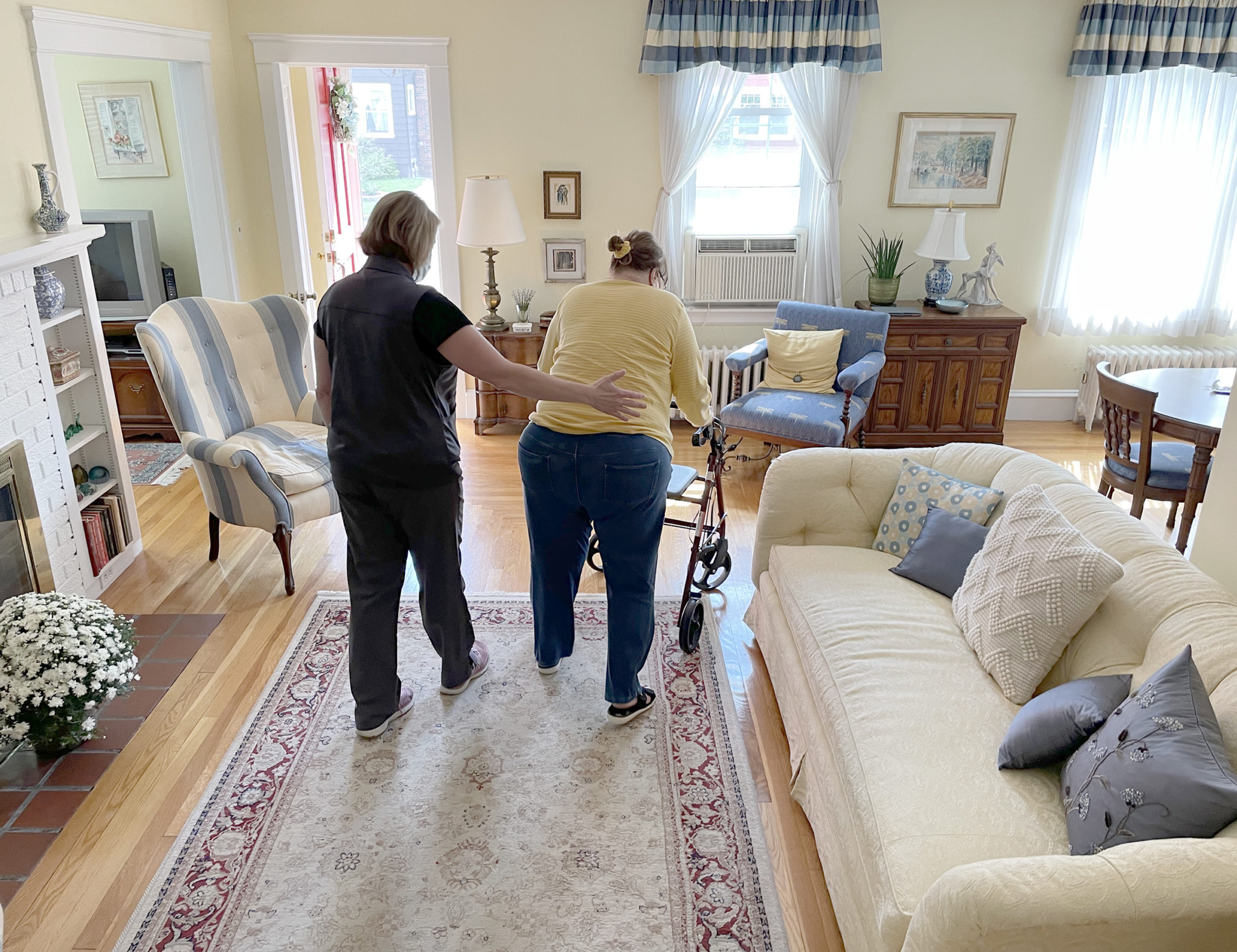 Physical therapy assistant walks behind older woman with walker in living room