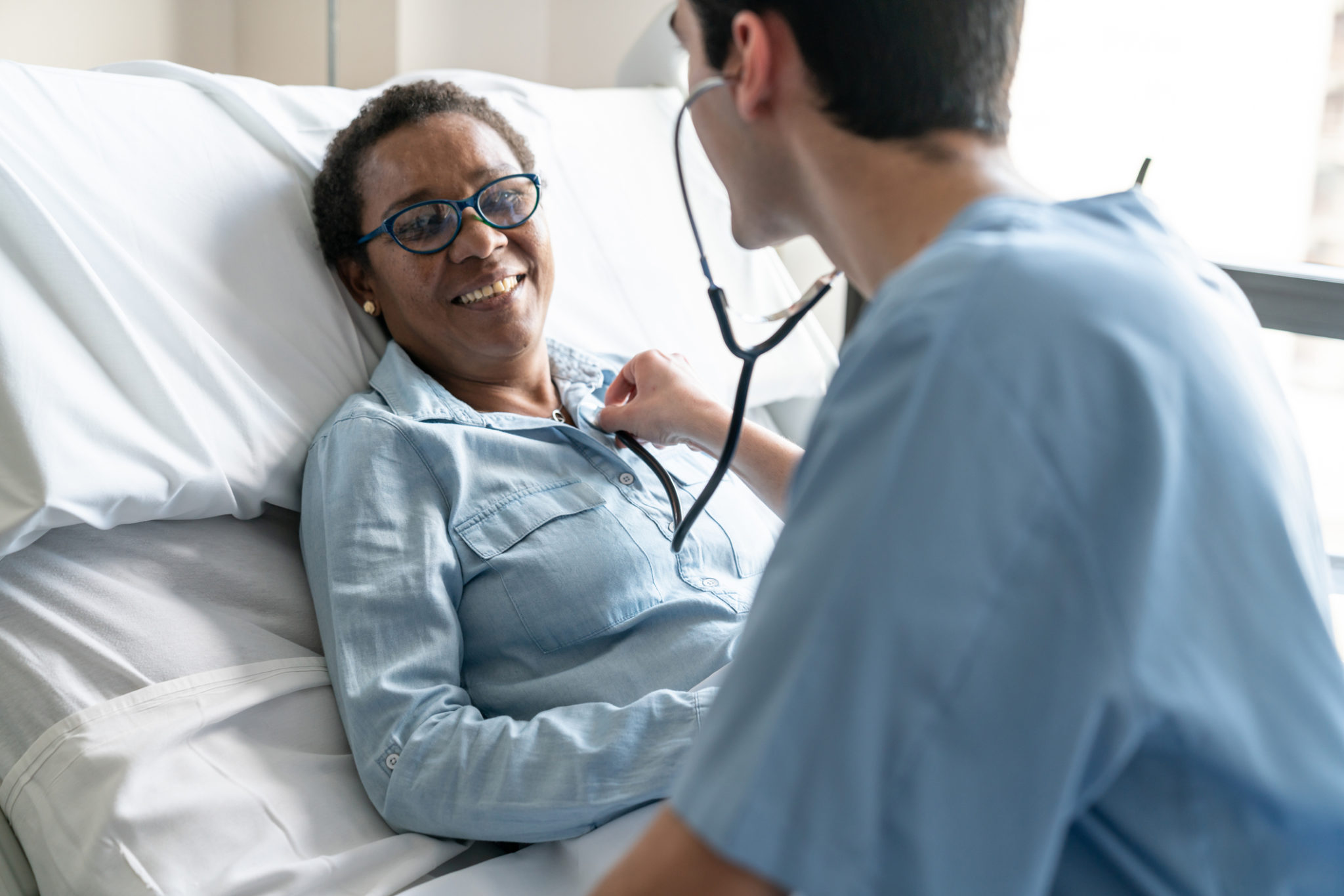 Female black patient lying down on hospital bed while nurse is checking her heart beat with stethoscope Female black friendly patient lying down on hospital bed while nurse is checking her heart beat with stethoscope