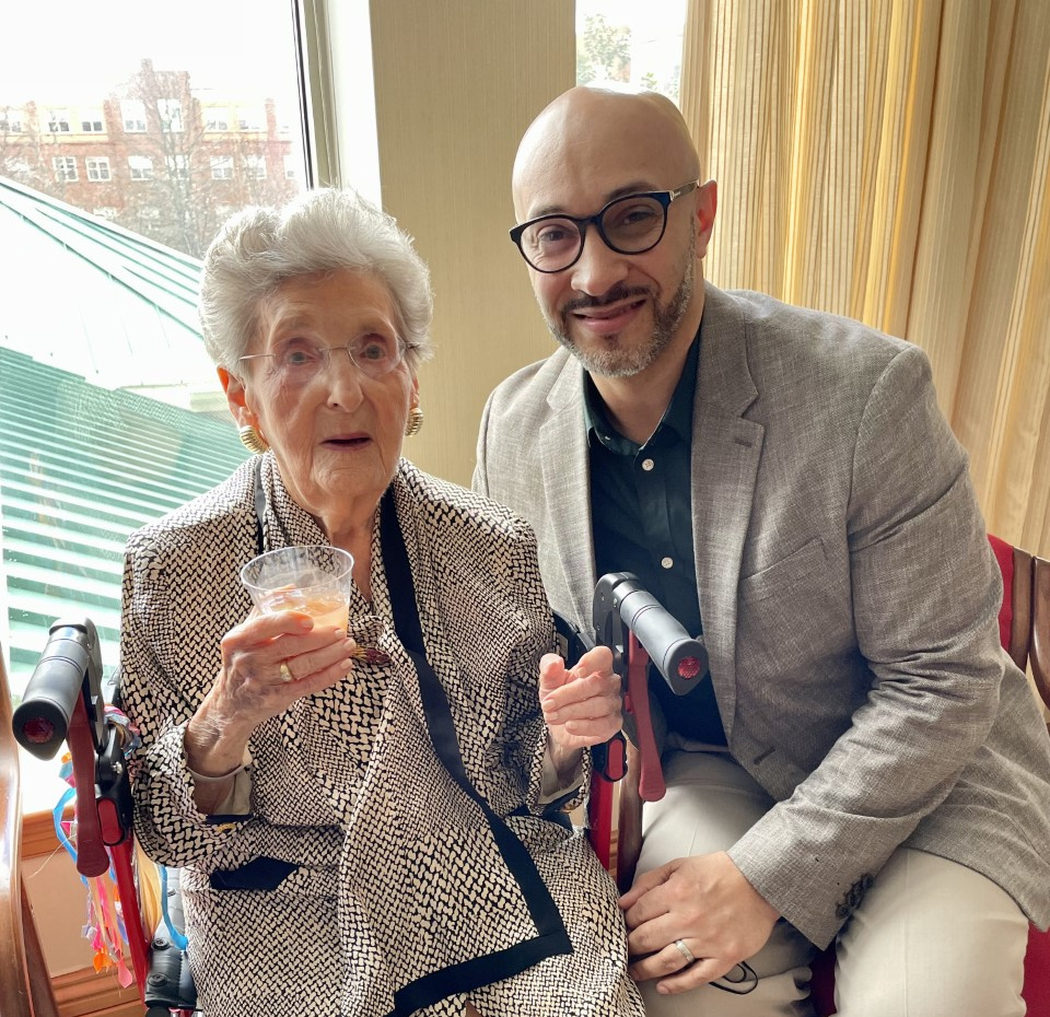 Elderly woman sitting with a drink in her hand as she poses with a younger man to her right