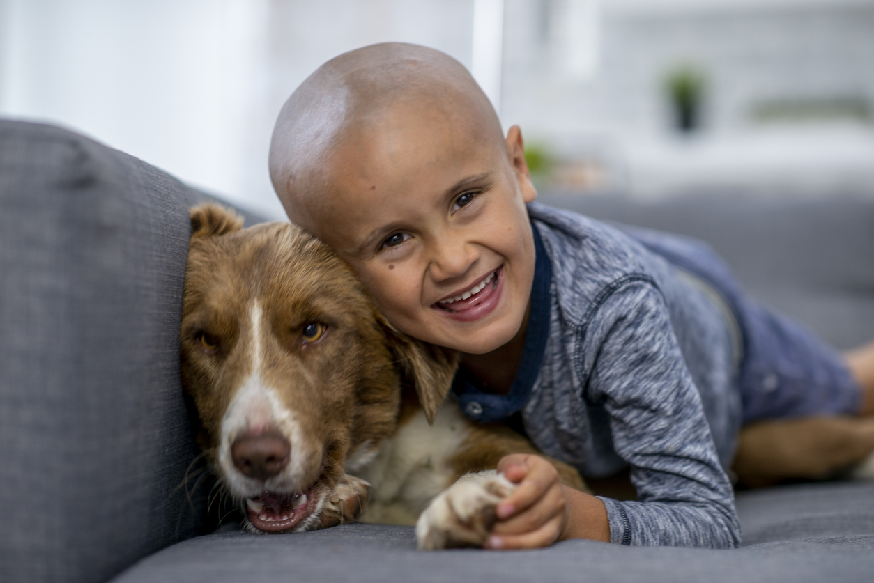 A young Ethnic boy is cuddling with his dog. He has cancer, and his head is shaved. He is happily smiling at the camera while lying on the couch.