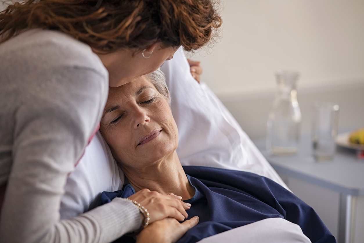 Close up of lovely daughter visiting and kissing sick mother on forehead while resting in hospital bed. Senior patient laying on gurney with eyes closed while daughter assuring a speedy recovery. Old mom patient being kissed while recovering from disease in nursing home.