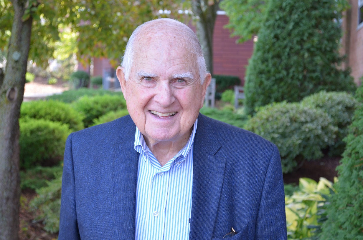 An older man smiles in a blue suit in front of greenery