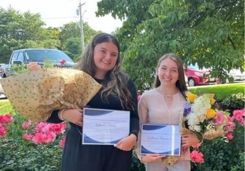 Two young girls standing in front of rose bushes with flowers and a graduation certificate