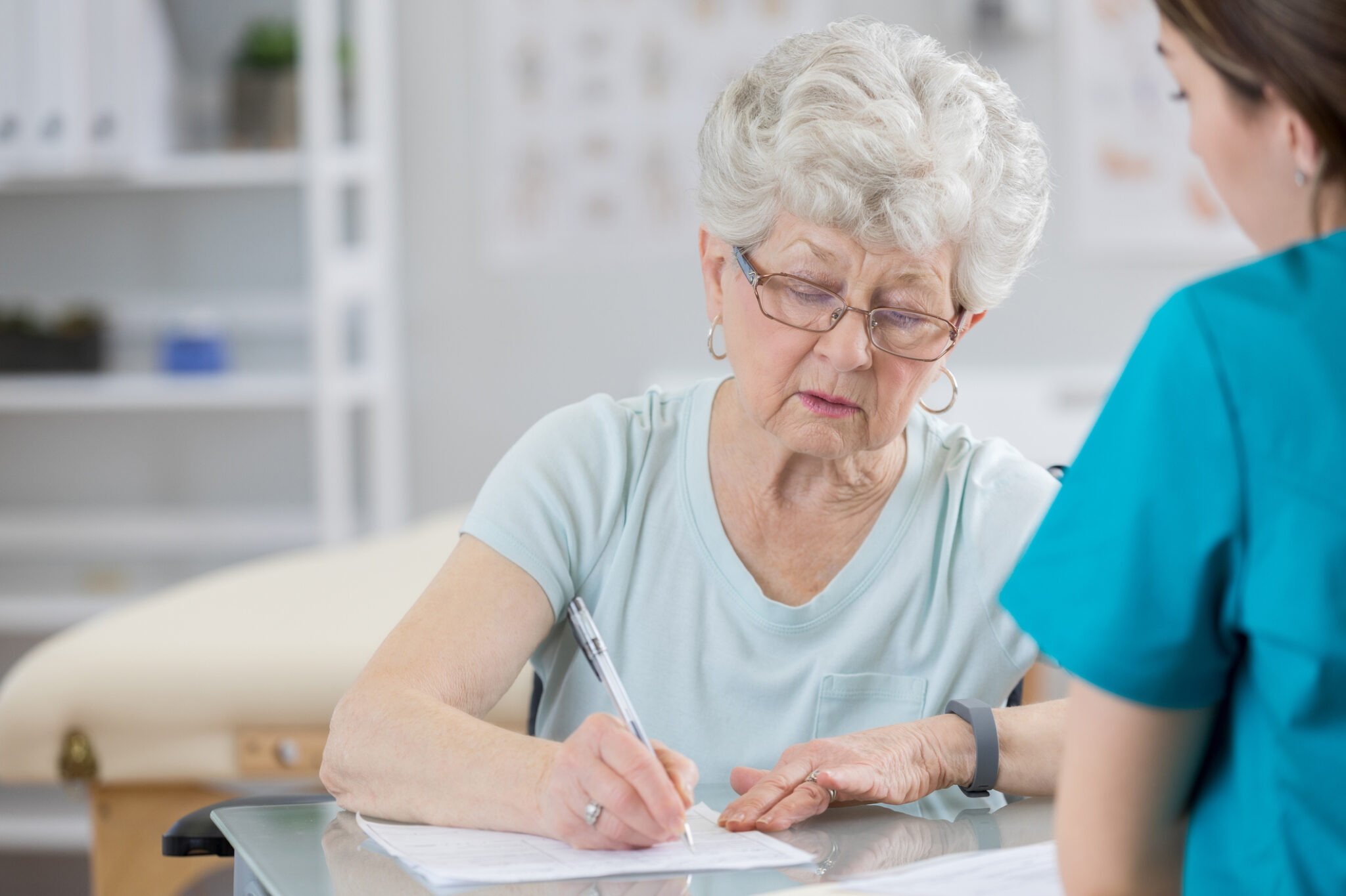 Senior woman concentrates as she completes insurance paperwork. A healthcare professional is helping her with the paperwork.