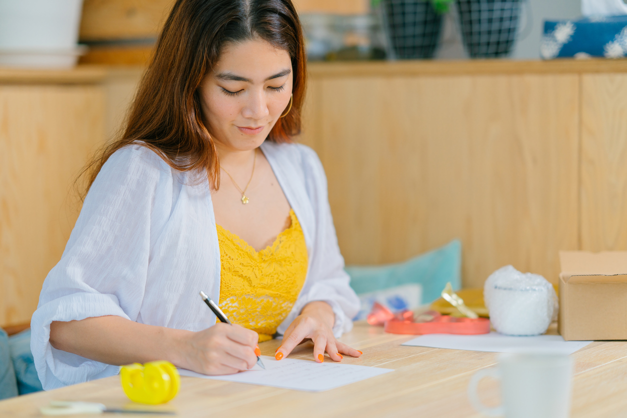 A young woman is writing a letter to put inside a gift box in the living room at home.