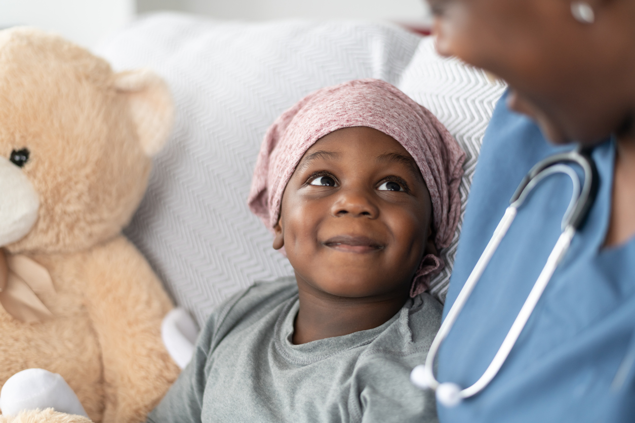 A young boy of African descent is at a medical consultation. He is dressed in casual clothes while wearing a headscarf to hide his hair loss. He sits beside his stuffed animal while listening to his doctor of African descent and smiling up at her.