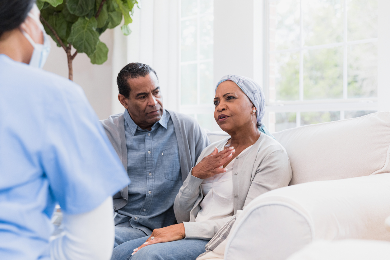 The senior adult husband watches worriedly as his wife describes her symptoms to the unrecognizable hospice nurse.