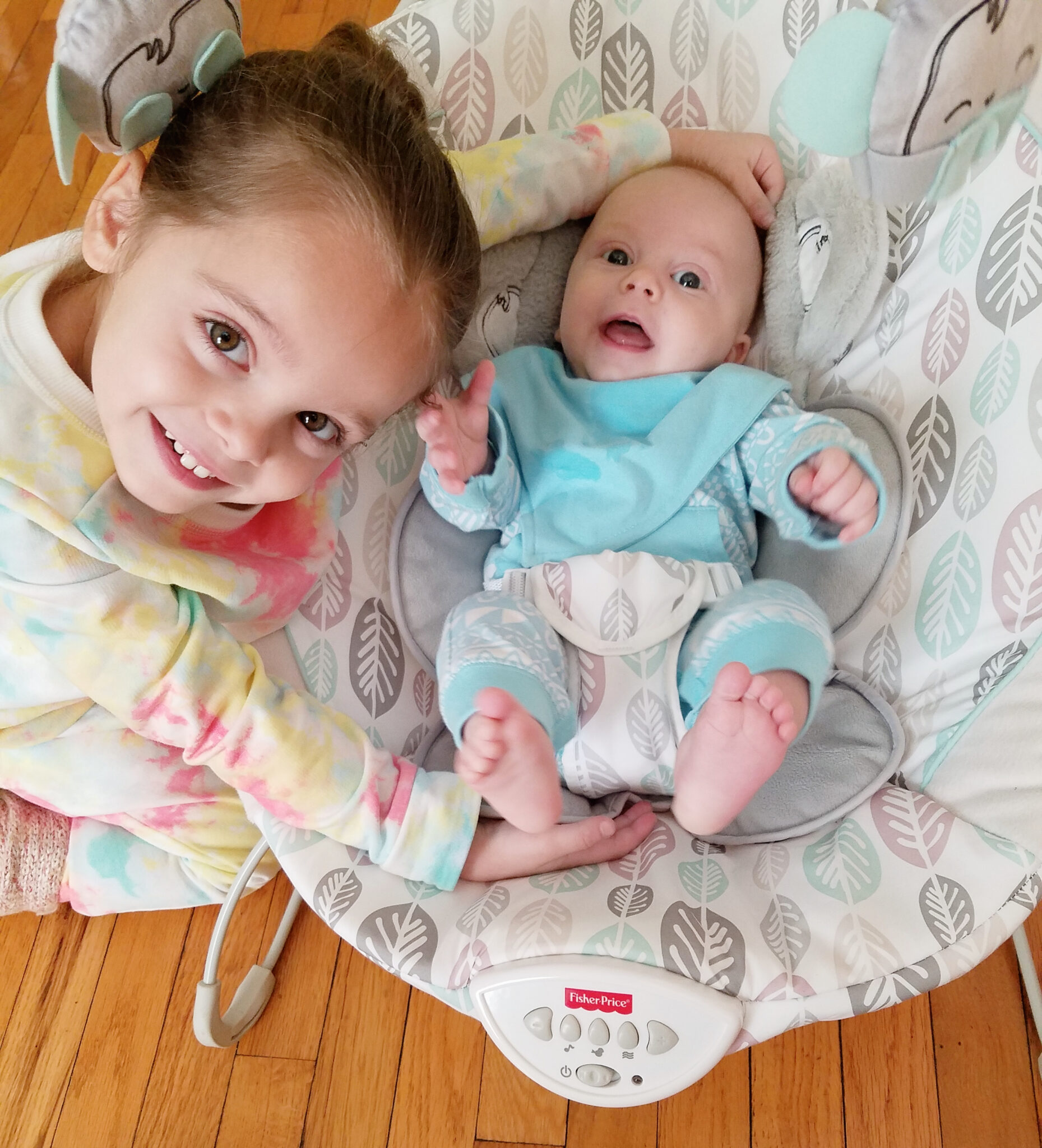 Baby boy smiling wide in a bouncy chair with his older sister leaning over him as she wraps her arm around his head