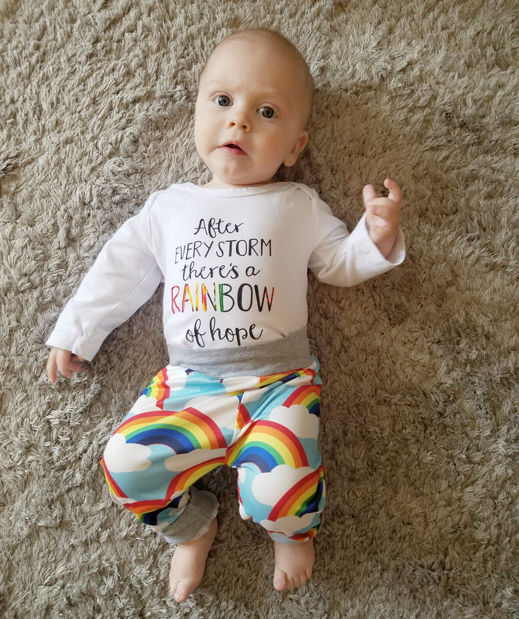 infant laying on the floor wearing rainbow pants and a white shirt that says, "after every storm there's a rainbow of hope"
