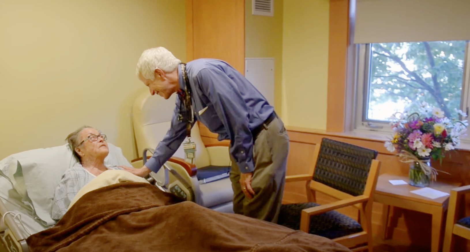 Doctor compassionately leans down to a patient in her bed, reaching out his arm in compassion