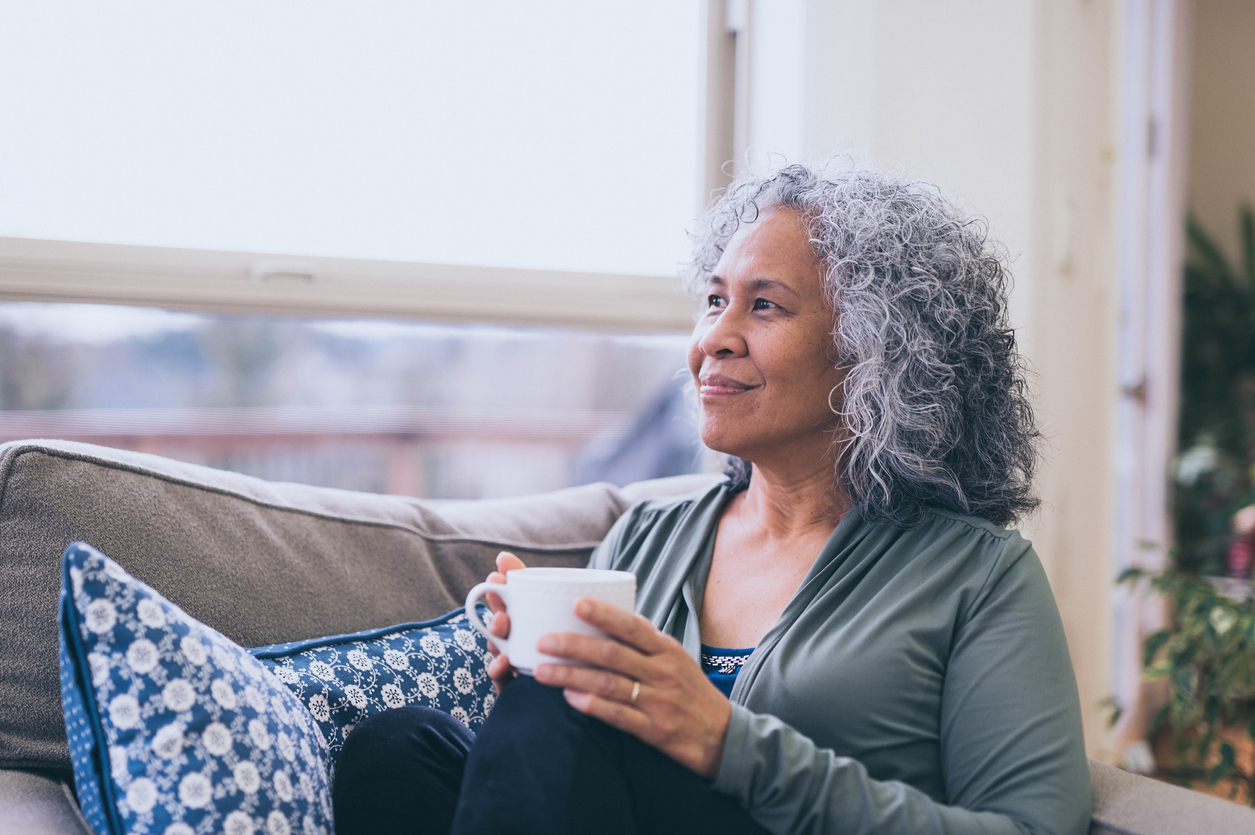 Woman with curly grey hair holding a white mug, sitting on the couch and looking out the window