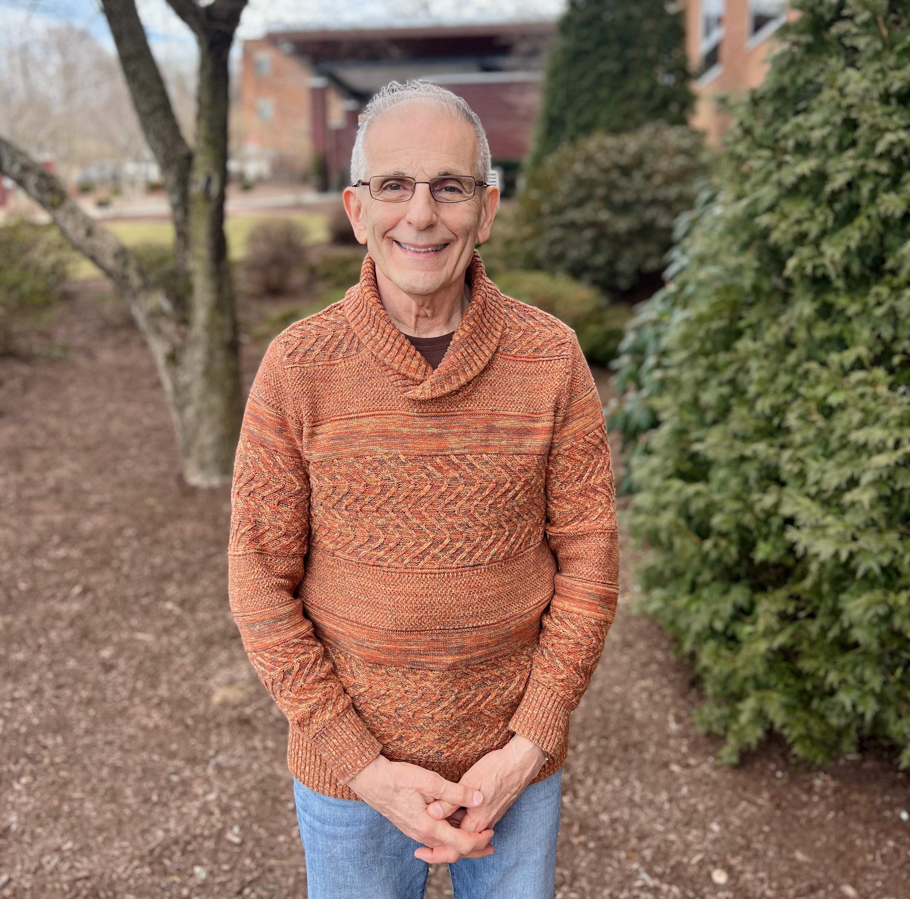 Man in burnt orange sweater wearing glasses stands in garden of the Hulitar Hospice Center with greenery surrounding him