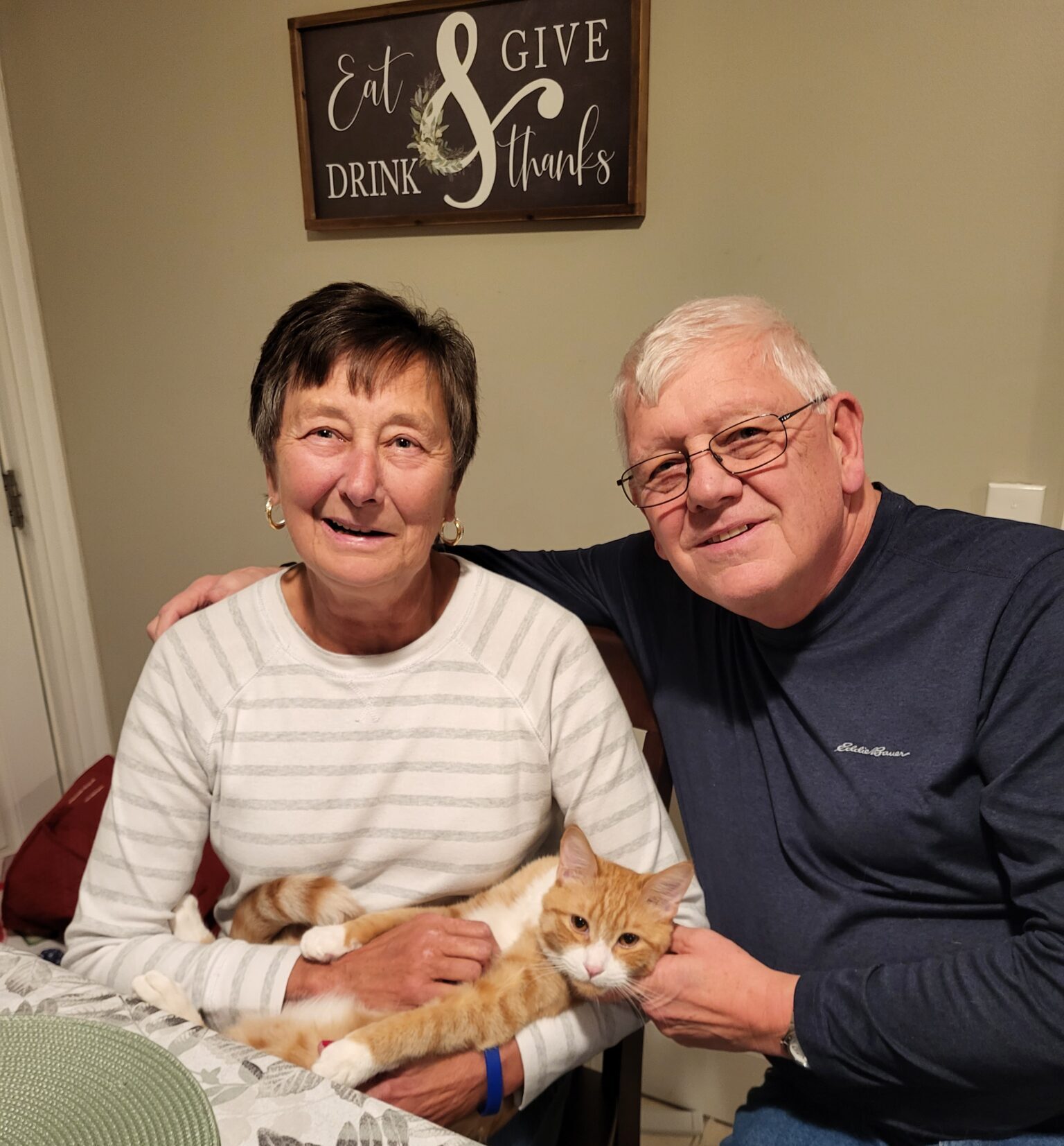 A man and women sit at the kitchen table. The man has his arm wrapped around the woman while she gently pats a cat.