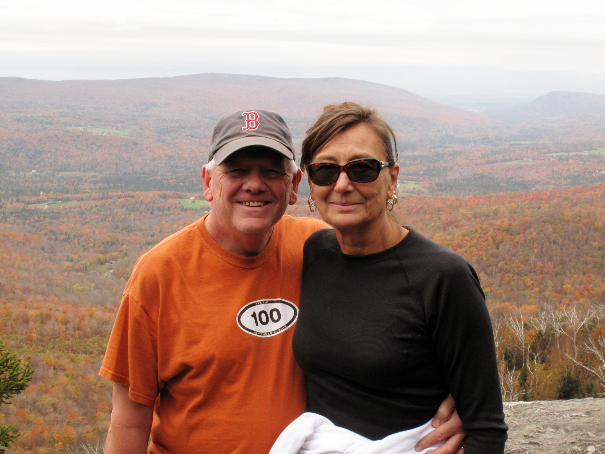 A man and woman embrace in front of a majestic outlook of orange foliage over a mountain range