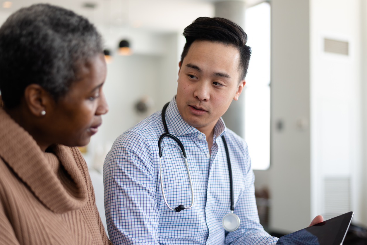 A doctor and an elderly patient are indoors at the woman's home. The doctor is smiling and talking to the woman while holding a tablet computer.