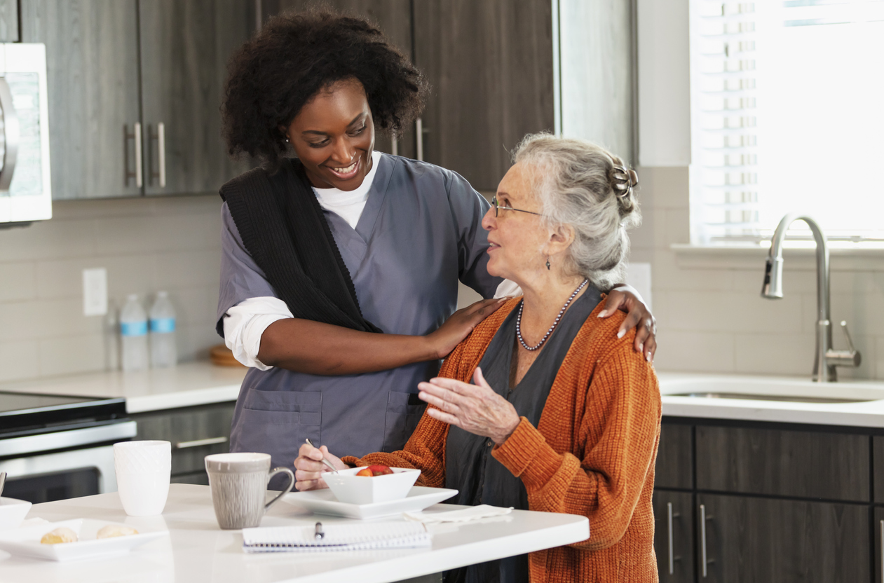 A home healthcare worker, an African-American woman in her 30s, helping a senior woman in her 80s eating a healthy snack in the kitchen. They are looking at each other, conversing.