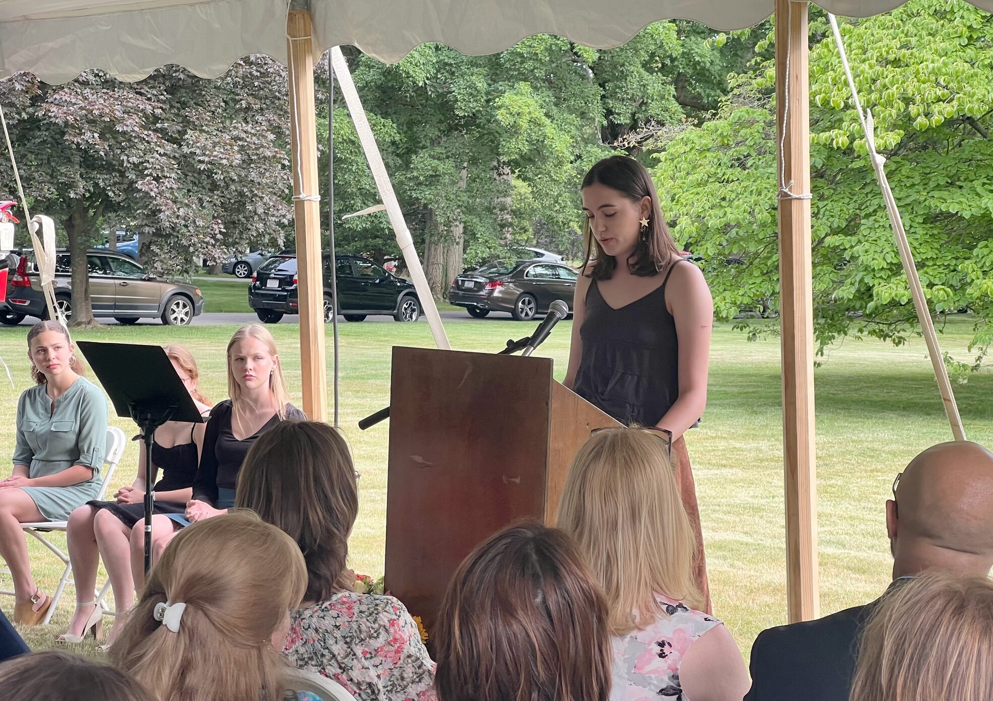 A young girl speaking at a podium under a tent to a group of seated people