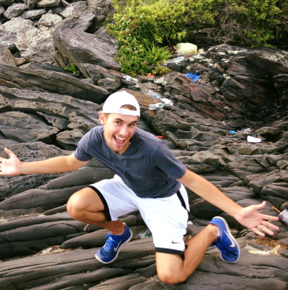 Young man with backwards white cap kneeling on ocean rocks with his hands stretched out