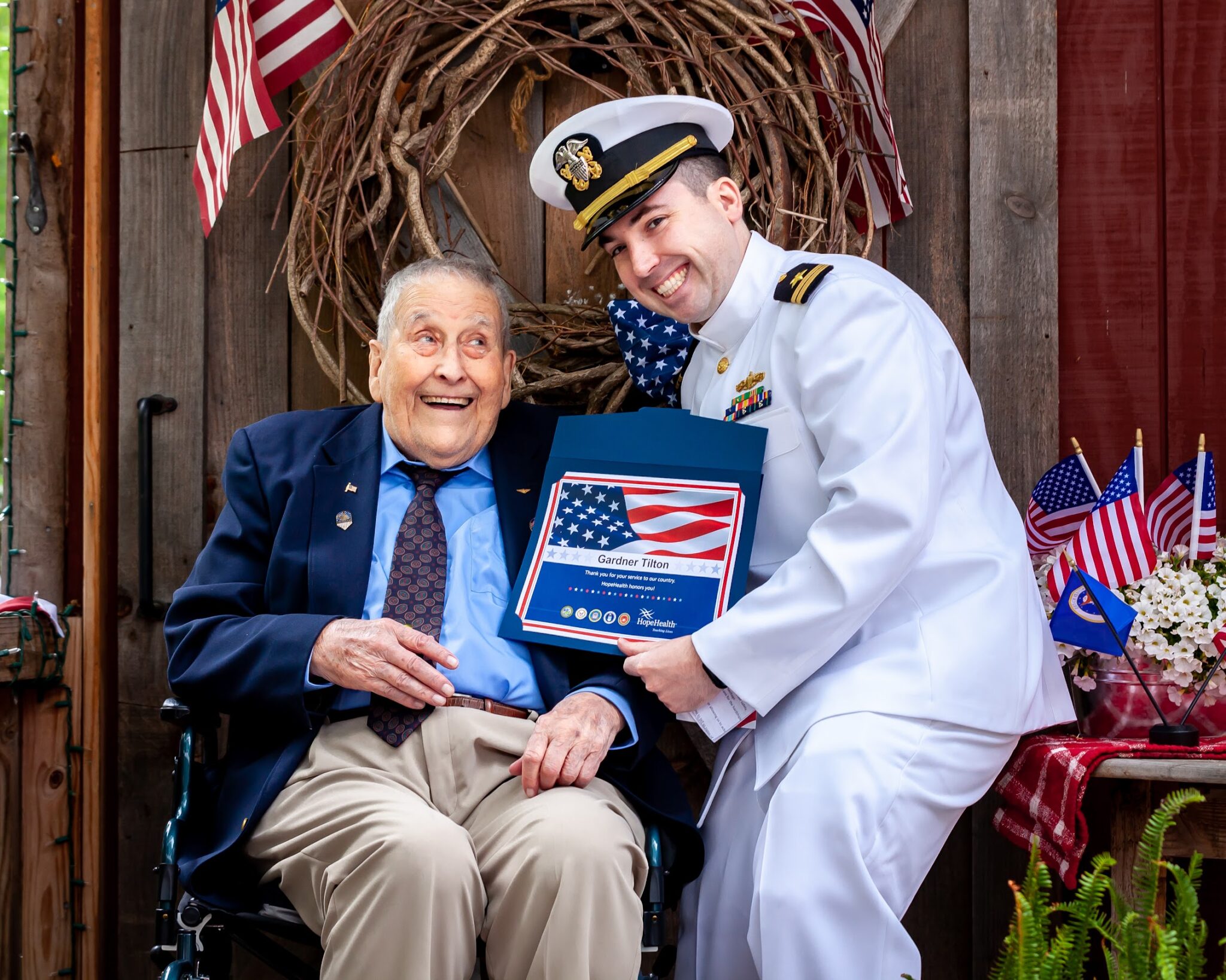 Major Gardner Tilton sits in his wheelchair in front of a patriotic flag wearing a navy blue suit jacket while receiving his pinning ceremony certificate from a HopeHealth volunteer in full navy uniform