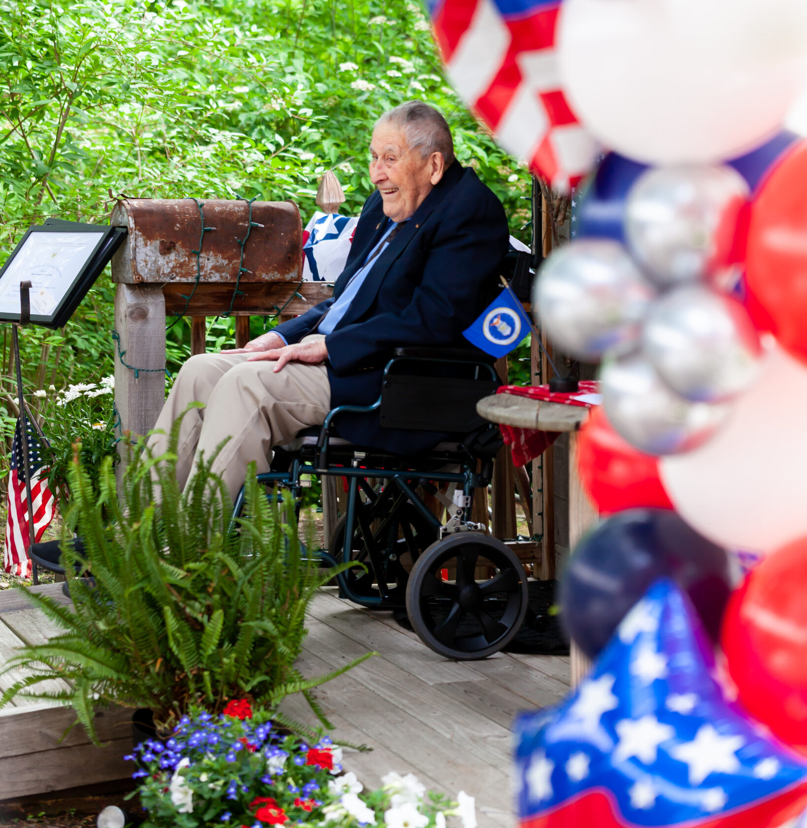 Veteran in navy suit jacket sits in a wheelchair surrounded by patriotic balloons