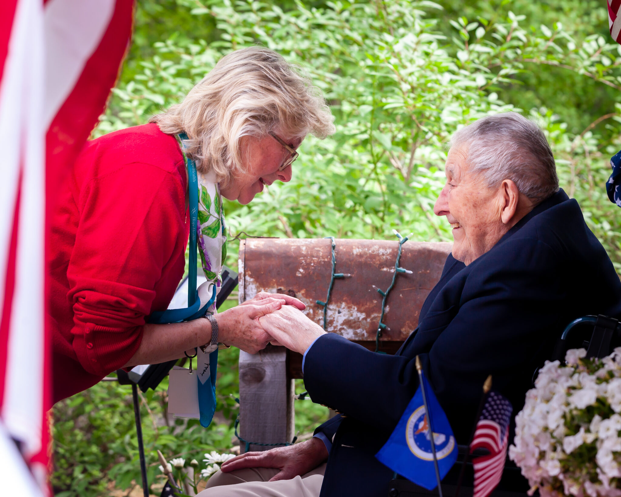 Woman in red sweater leans over to hold hands with an elderly veteran sitting in a wheelchair wearing a navy blazer