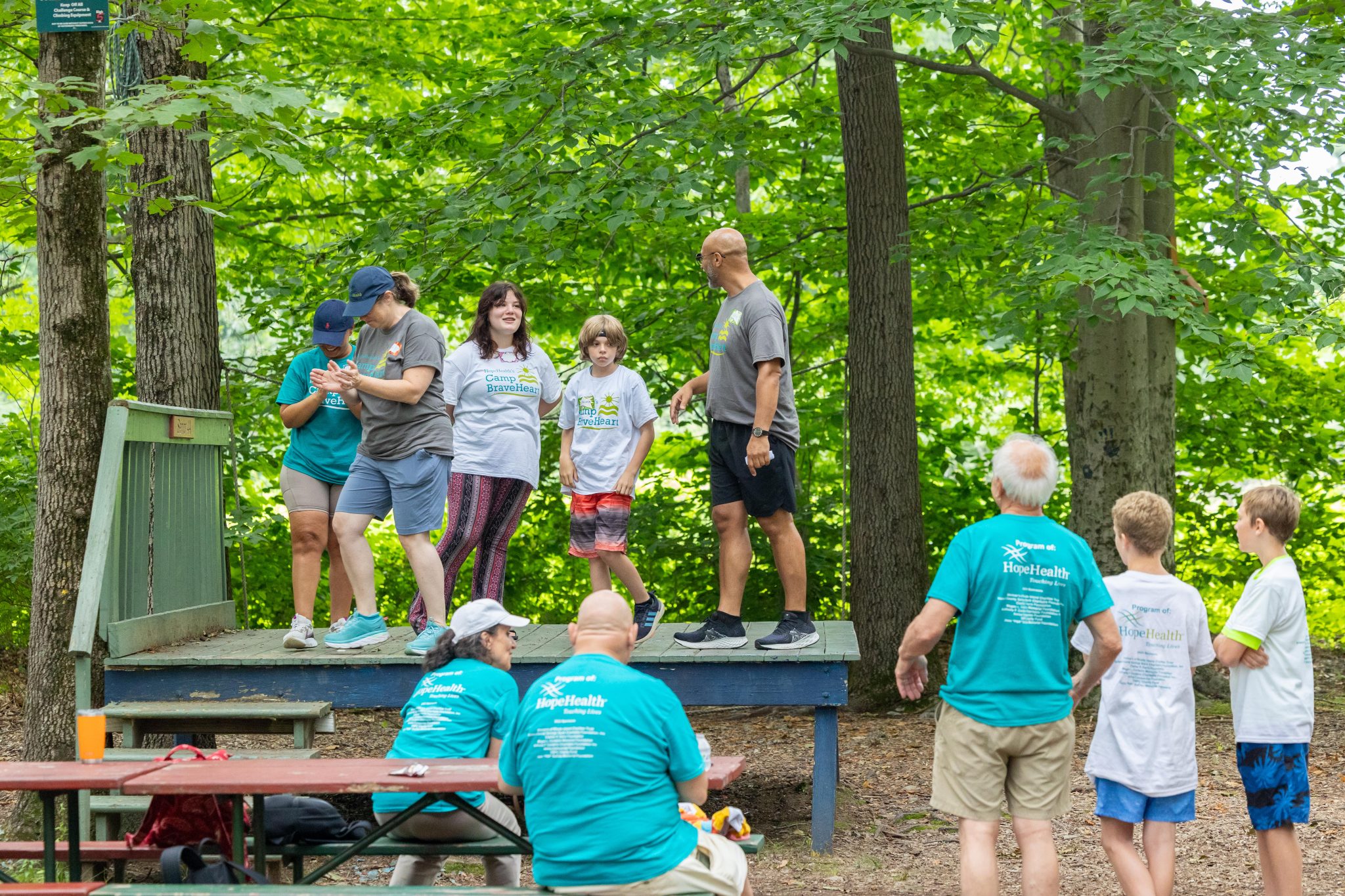 A group of kids stand on a theater stage in the woods while others look on