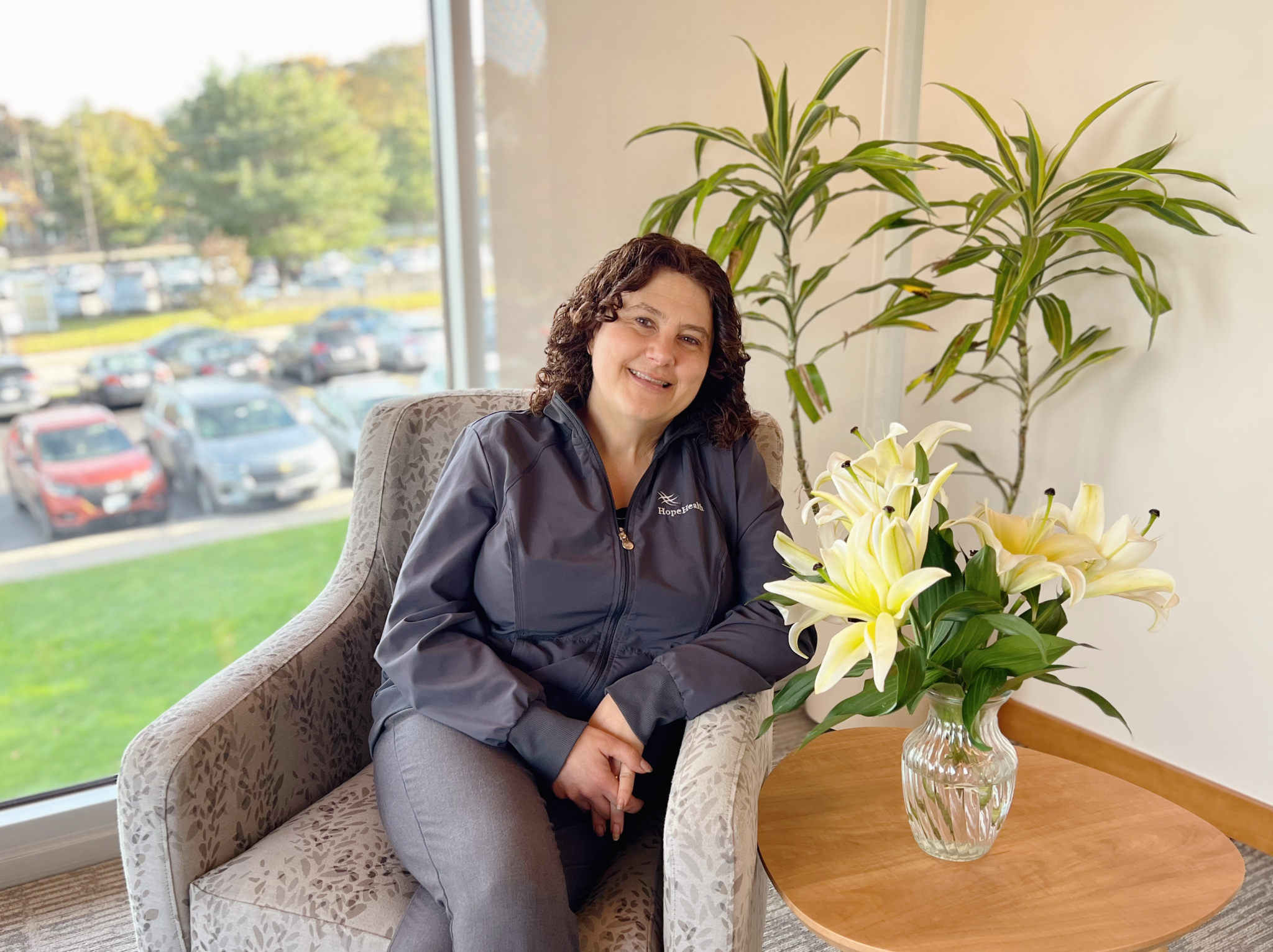 female palliative social worker with brown curly hair sitting in a arm chair next to a vase of white lillies