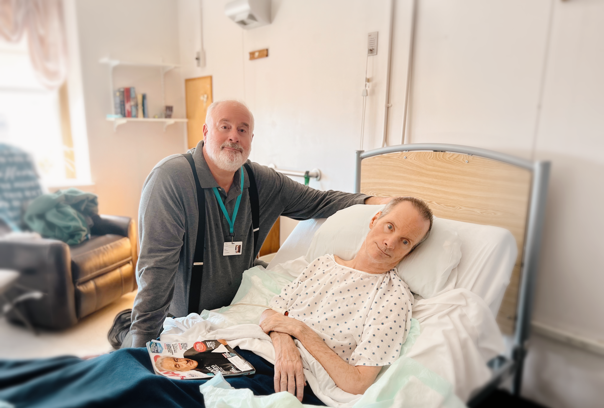 A man in a gray shirt and suspenders compassionately sits by a male hospice patient who is laying in a hospital bed