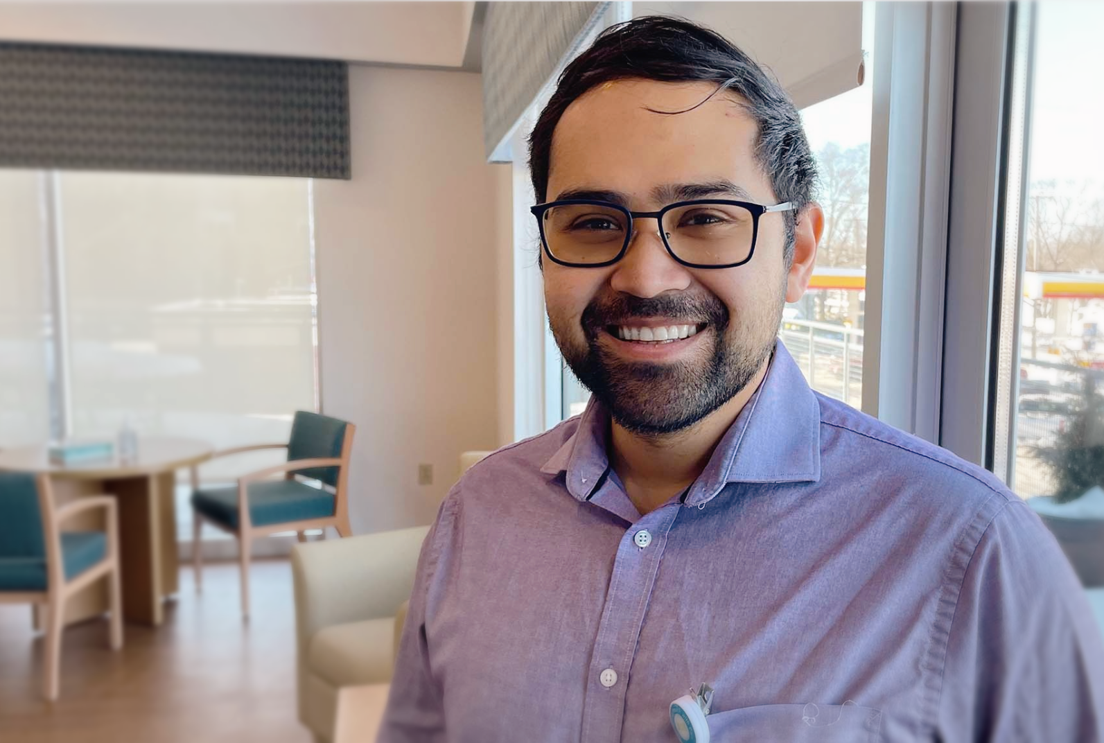 Dr. Jacob Ramos, a hospice physician, smiling warmly while standing in a sunlit patient consultation room with modern furnishings and large windows.