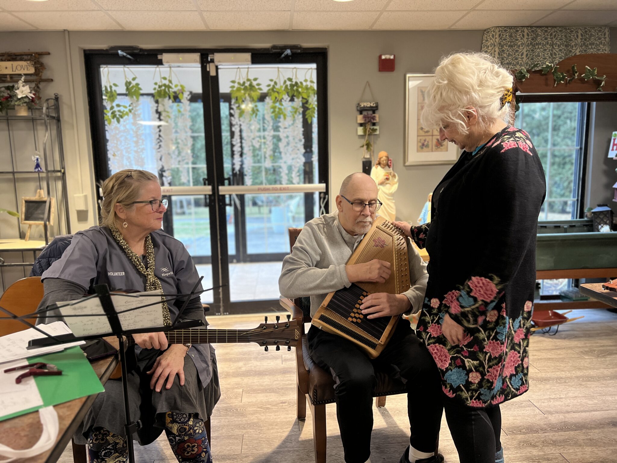 A volunteer in a gray smock holds a guitar, looking at a dementia patient holding a auto-harp while his wife stands next to him