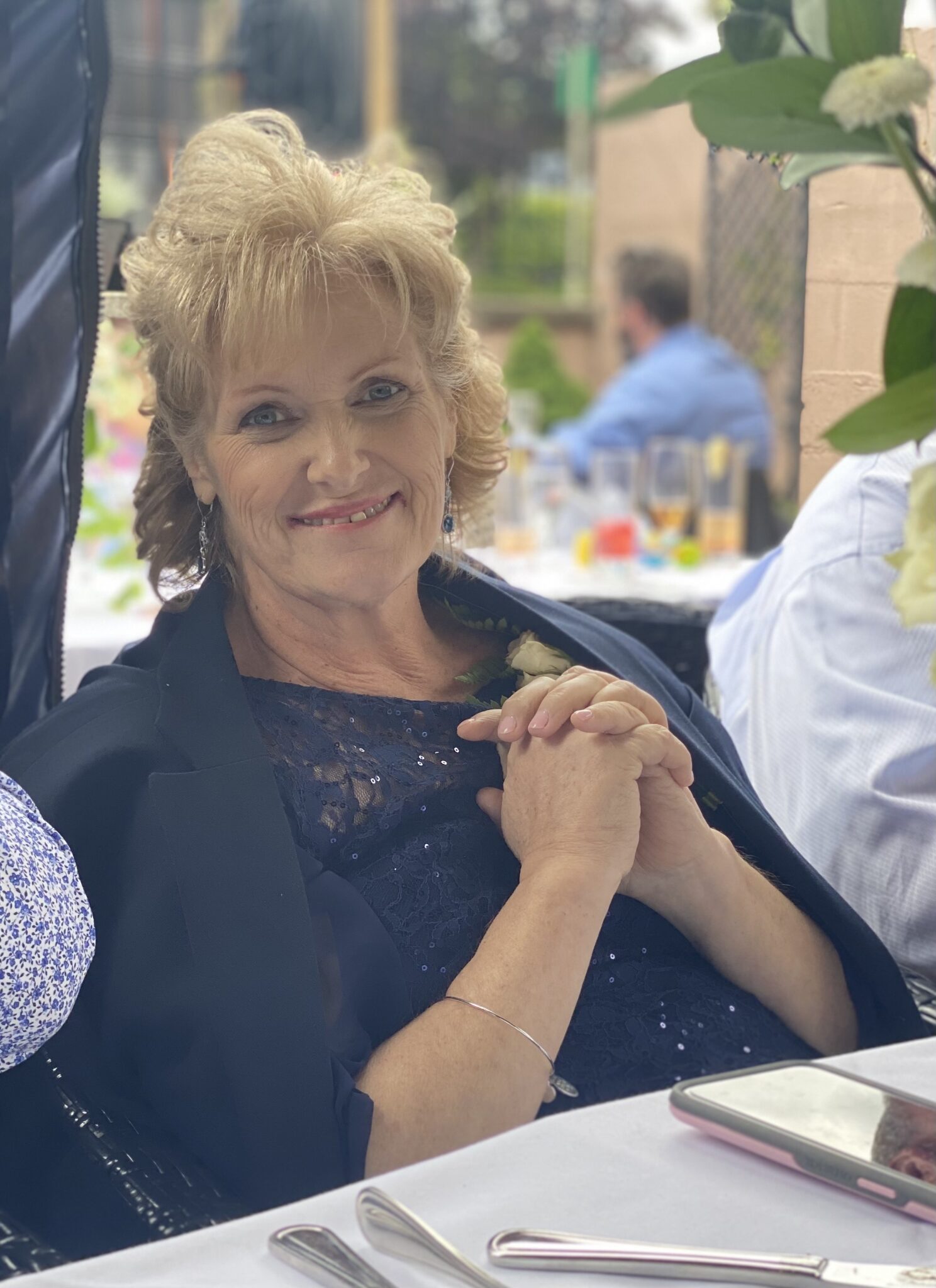 Blonde woman sitting at a table wearing a navy blue dress, clasping her hands in front of her with a bright smile