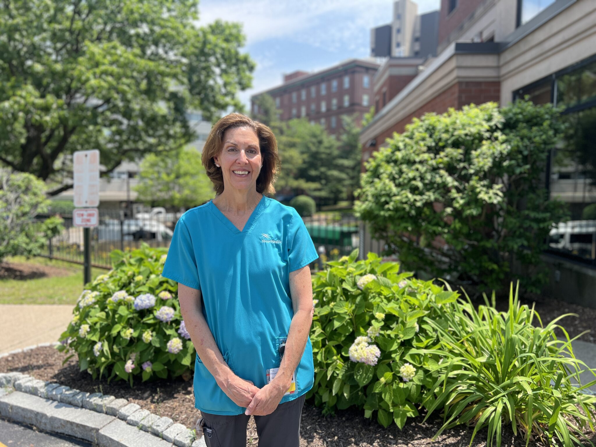 Female hospice aide standing in front of hydrangea bushes and brick buildings