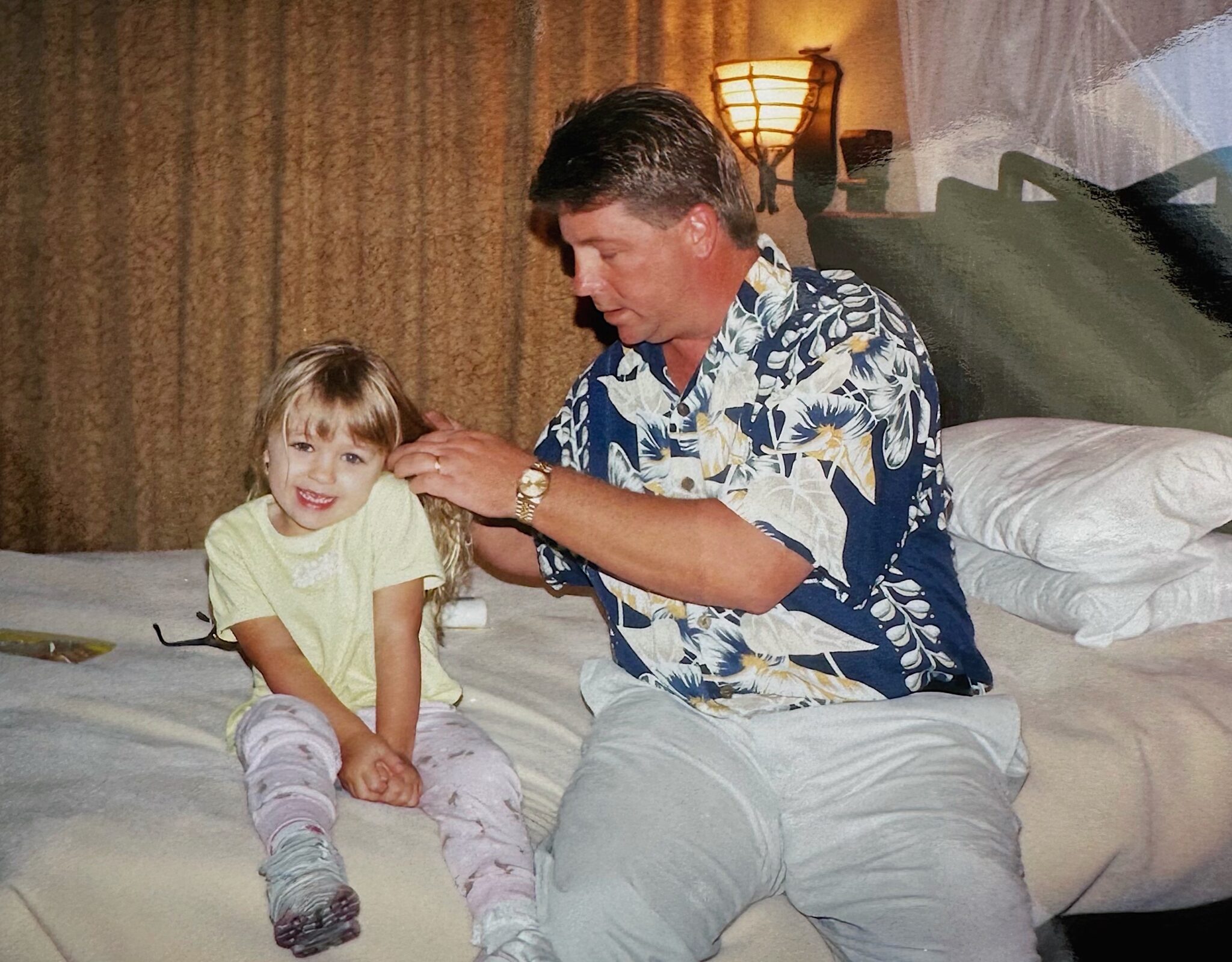 A man sits on a bed gently brushing his young daughter's hair. The girl smiles at the camera while the man focuses on her hair, and they are in a warmly lit bedroom.