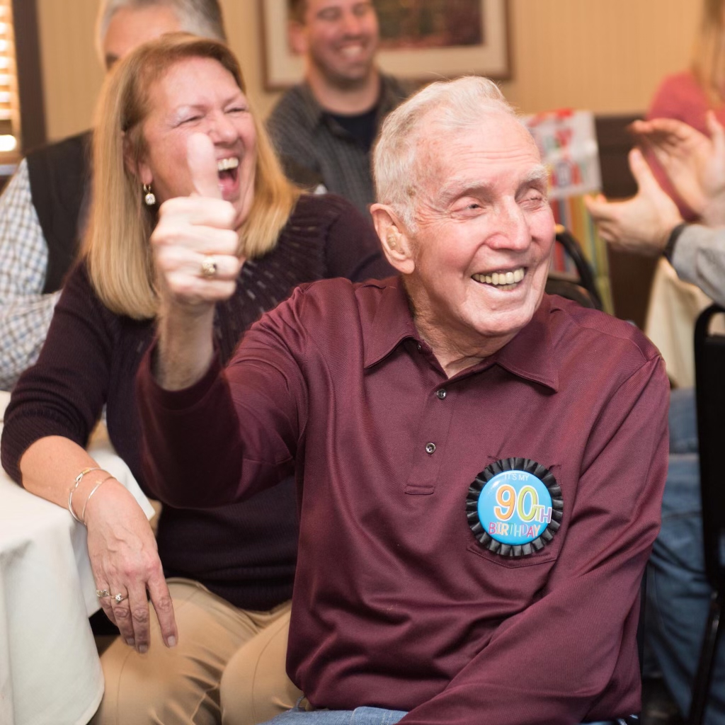 An elderly man celebrating his 90th birthday smiles broadly and gives a thumbs-up while sitting at a table surrounded by friends and family. He is wearing a maroon shirt with a “90th Birthday” badge on his chest. The atmosphere is joyful, with people laughing and clapping in the background.