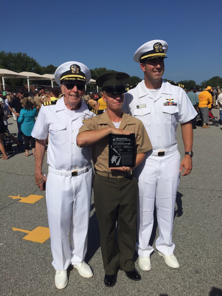 Three men in military uniforms stand proudly outdoors during a ceremony. The man in the center, dressed in a Marine uniform, holds a plaque while flanked by two men in white Navy uniforms.
