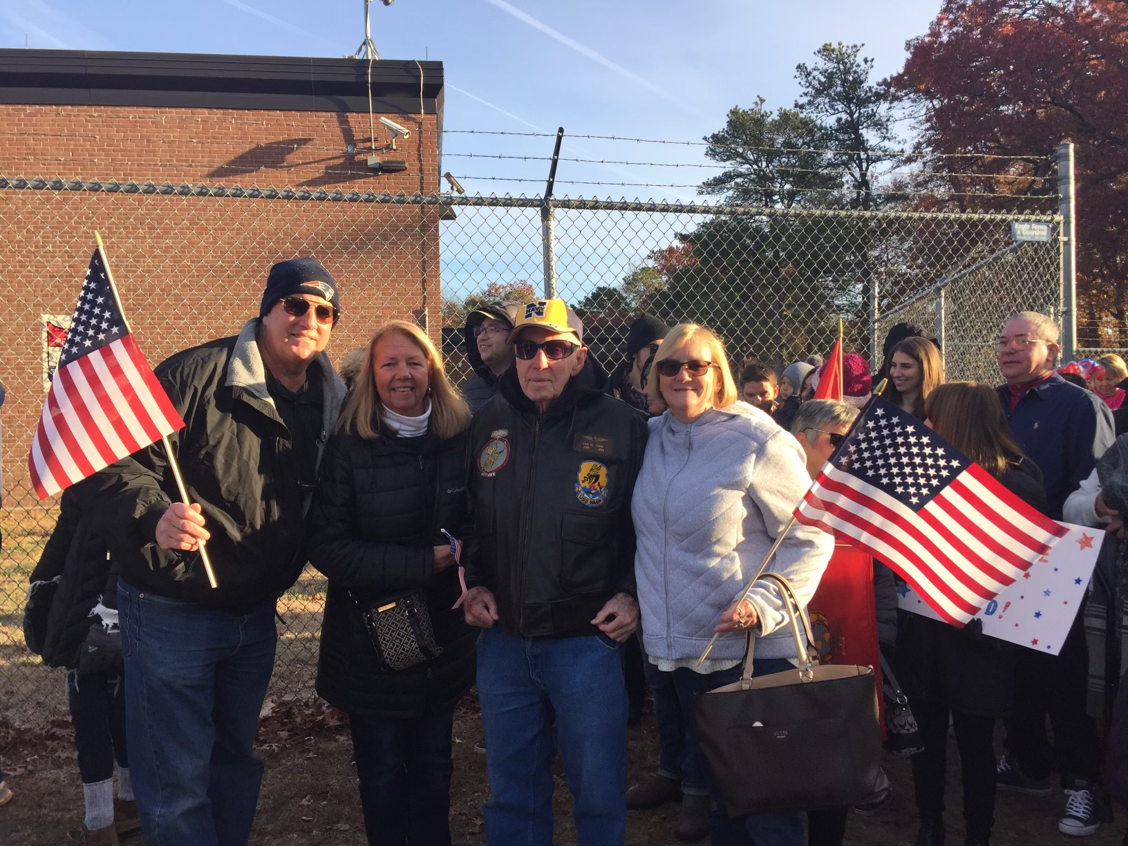A group of people gathers outdoors near a chain-link fence on a sunny day. They hold American flags and smile, with an older man in a leather jacket and hat standing proudly in the middle.