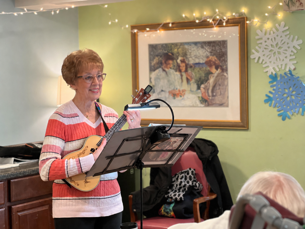 An older woman in a striped sweater smiles while playing a ukulele in a softly lit room, with a music stand in front of her and decorations on the wall behind.