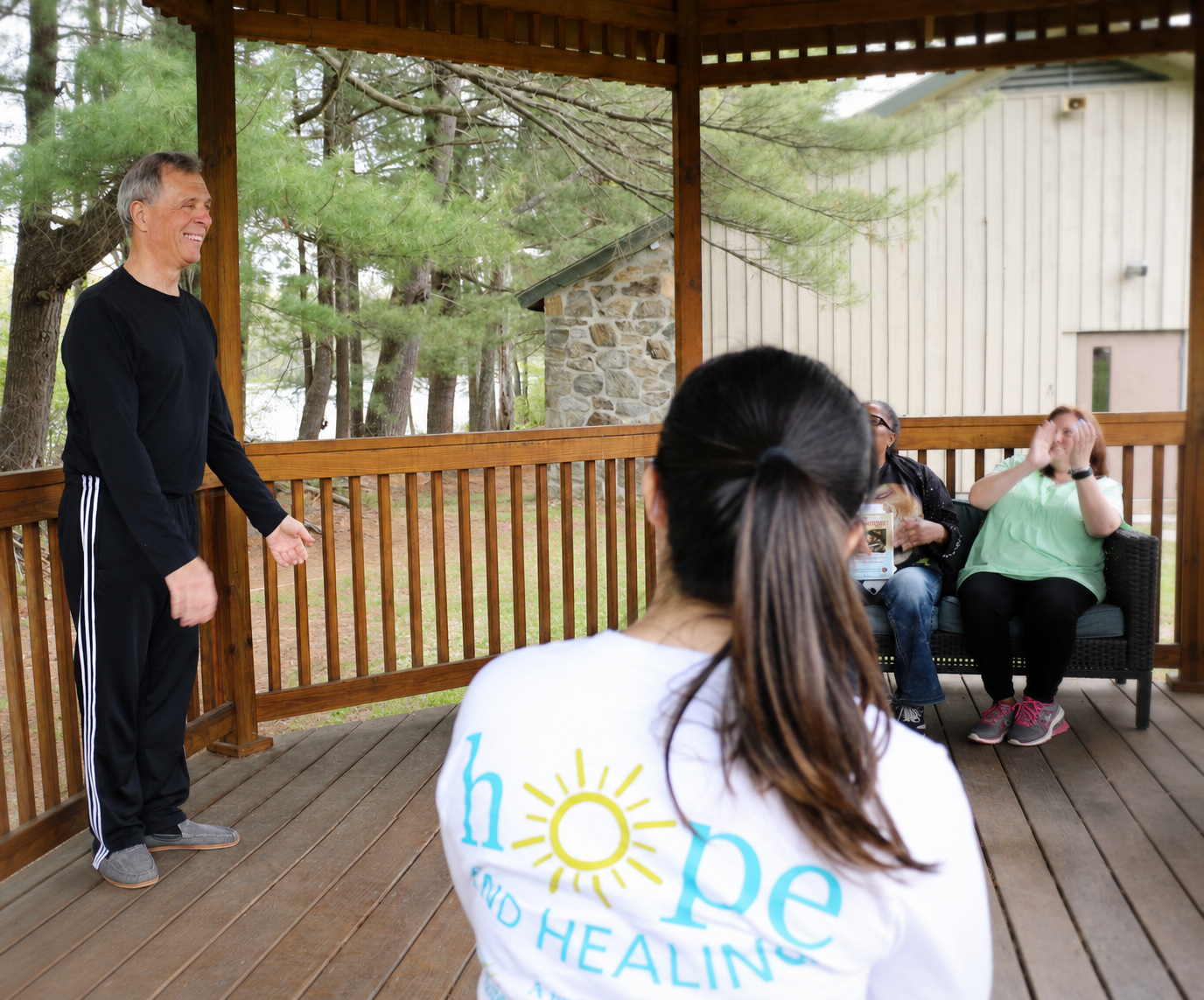Retired grief counselor Guy Murgo smiles while leading a group session during the 2017 Weekend of Hope & Healing. He stands on a covered wooden pavilion surrounded by pine trees as participants listen and applaud, creating a warm and supportive outdoor gathering atmosphere.