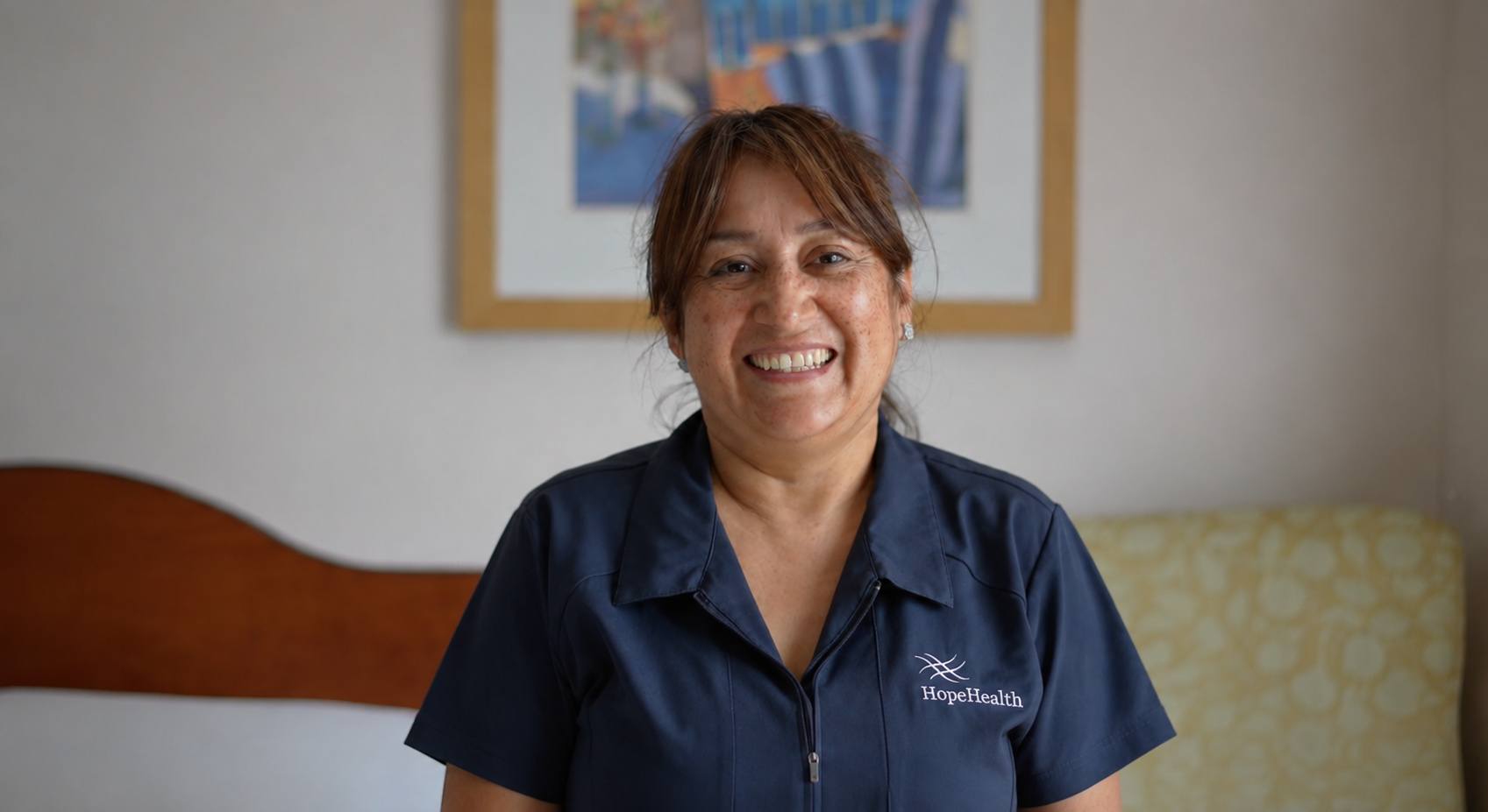 Nora Navas, CNA, stands in a patient room at the HopeHealth Huliar Hospice Center wearing gray scrubs, smiling warmly