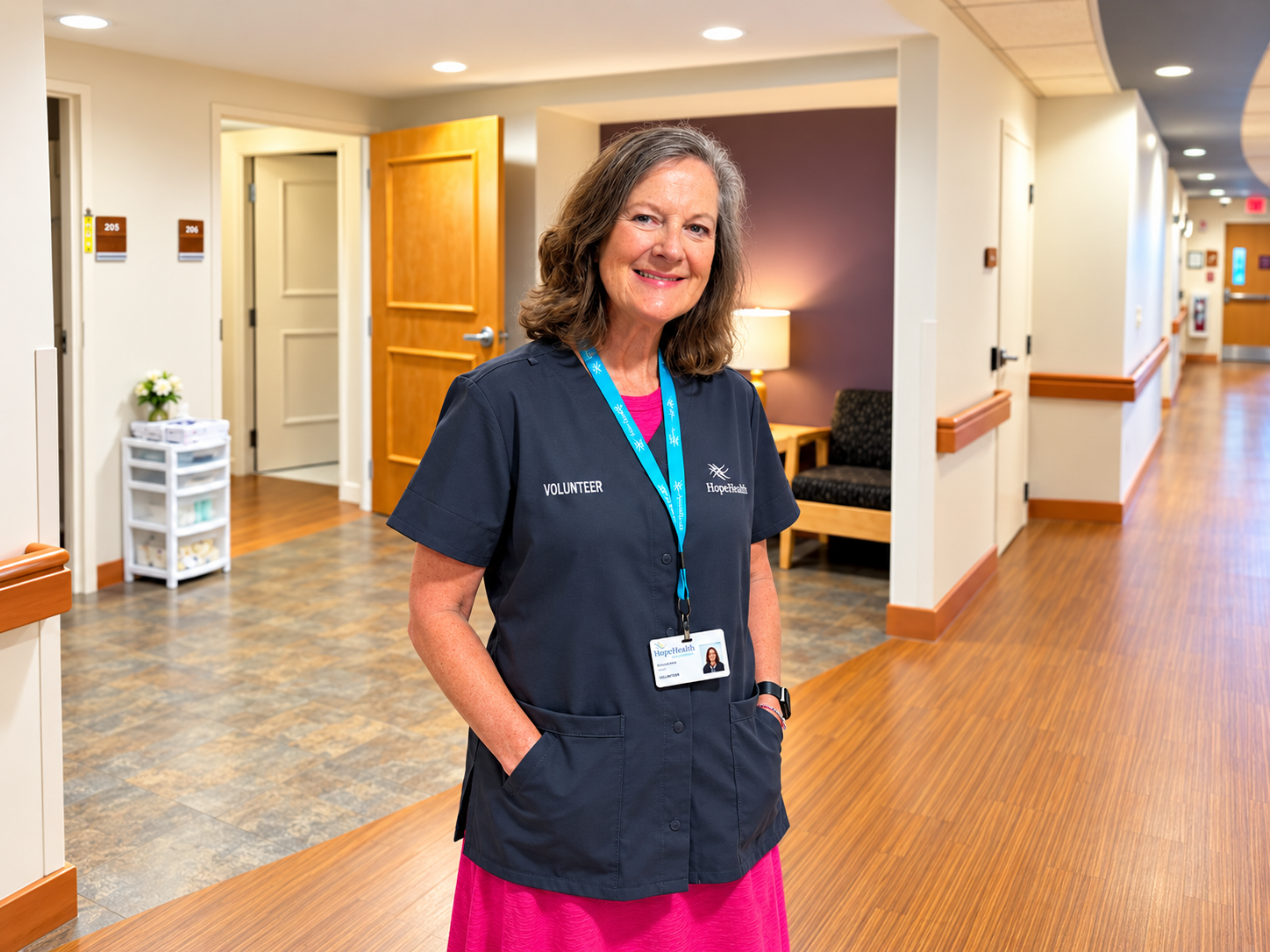 Woman in pink dress and gray volunteer smock smils while standing in front of hospice IPU rooms