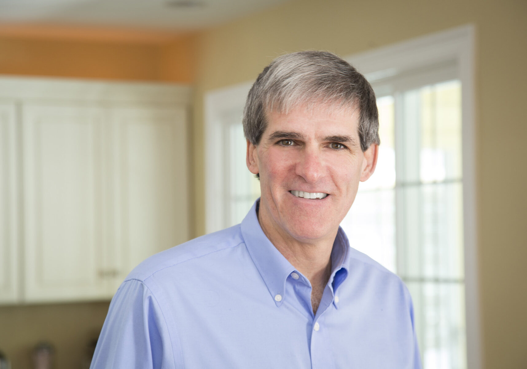 Older man wearing light blue button down shirt smiling in his kitchen