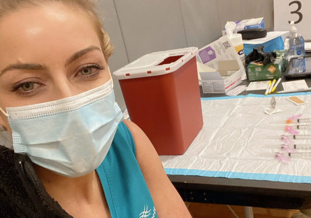 Female nurse wearing teal scrubs smiles as gets COVID-19 vaccine