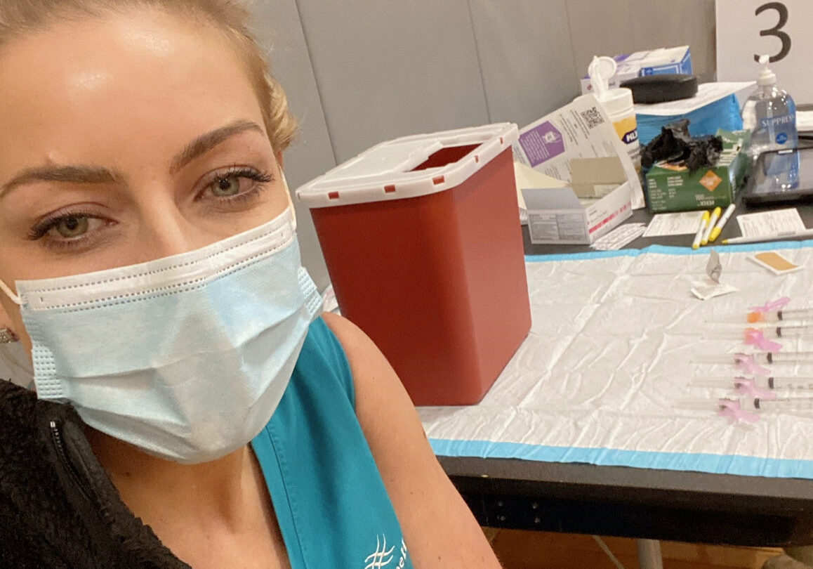 Female nurse wearing teal scrubs smiles as gets COVID-19 vaccine