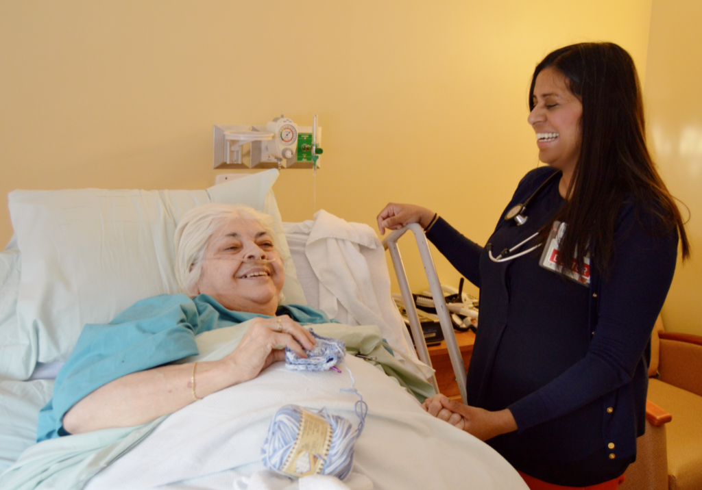 A woman laying in bed and knitting while she laughs and talks to her hospice doctor. She is receiving inpatient hospice care at the Hulitar Hospice Care Center