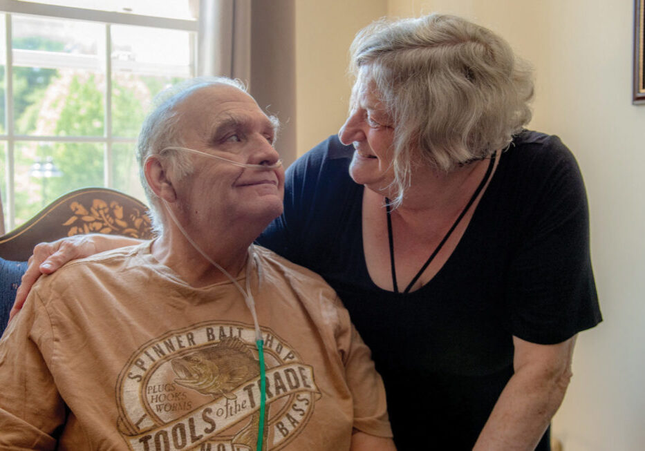 An elderly couple looks lovingly at each other. The man is sitting and wearing an oxygen tank while the woman is standing next to him with her arm around him.