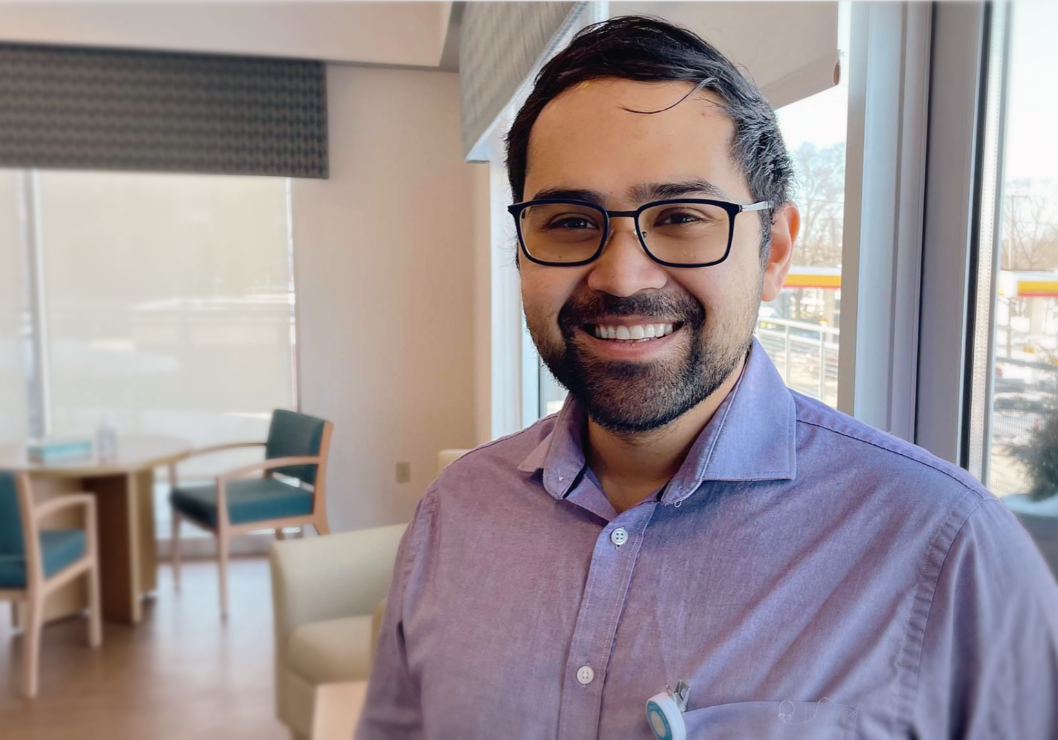 Dr. Jacob Ramos, a hospice physician, smiling warmly while standing in a sunlit patient consultation room with modern furnishings and large windows.