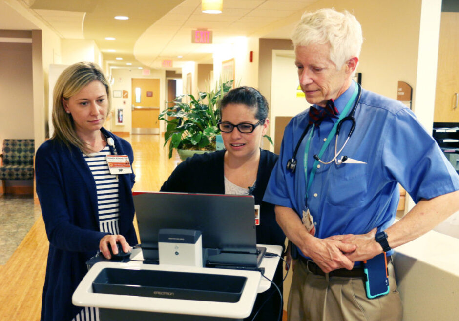 Two young women stand with an older man while reviewing a laptop in front of them