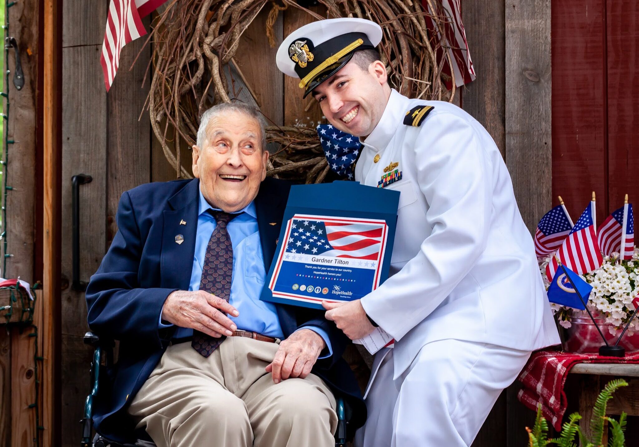 Major Gardner Tilton sits in his wheelchair in front of a patriotic flag wearing a navy blue suit jacket while receiving his pinning ceremony certificate from a HopeHealth volunteer in full navy uniform